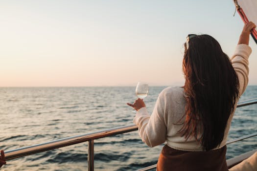 A woman with long hair holds a drink on a sailboat, enjoying a sunset view at sea.